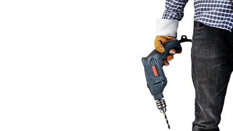 Maschinen sollen geeignet und sicher sein In der rechten Bildhälfte sieht man die Körpermitte eines Mannes, der Arbeitshandschuhe trägt und eine Bohrmaschine in der Hand hält