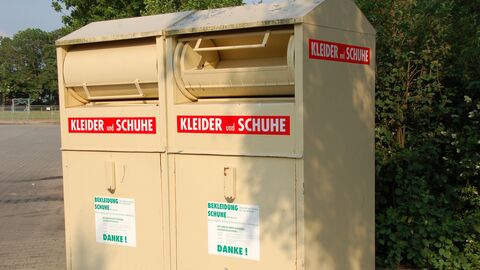 Ein beiger Altkleidercontainer mit der Aufschrift Kleider und Schuhe steht vor grünen Büschen 