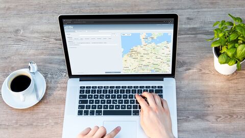 Zwei Hände tippen am Laptop, auf dem eine Landkarte zu sehen ist. Links neben dem Laptop steht eine Kaffeetasse und rechts eine Basilikumpflanze. 