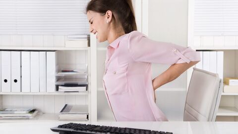 Der Betriebsarzt macht dem Arbeitgeber Vorschläge zur Verhütung arbeitsbedingter Erkrankungen Junge Frau sitzt am Schreibtisch und greift sich schmerzverzerrt in den Rücken