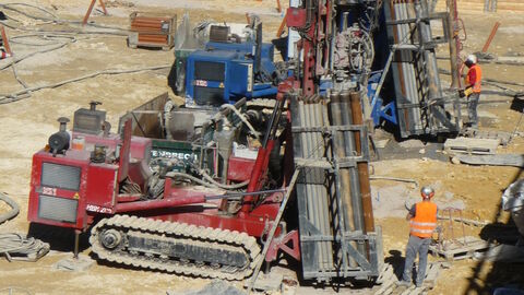 Zwei Männer, mit orangen Warnwesten, stehen neben großen Bohrgeräten auf gelbem, sandigen Boden. 