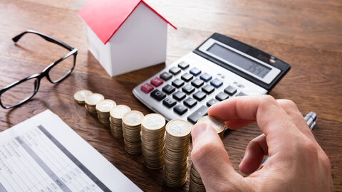 Auf einem Tisch sieht man eine Berechnung, einen Taschenrechner, eine Brille und eine Hand, die gestapeltes Kleingeld zählt