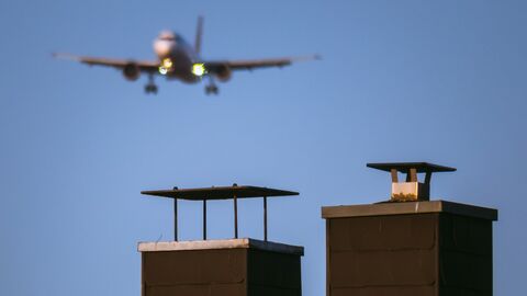 Nahaufnahme eines Gebäudedachs mit Schornstein über dem im Hintergrund ein Flugzeug zu erkennen ist