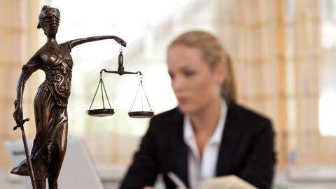 Recht und Gesetz Bronzestatue der Justitia, dahinter eine Frau am Schreibtisch mit Büchern und Laptop