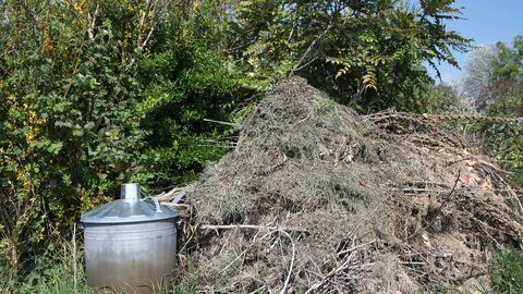 Viele Äste und Grünschnitt liegen auf einem Haufen im Freien, links daneben steht eine silberne Tonne mit Deckel
