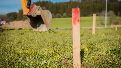 Holzpflöcke auf einer Wiese stecken ein Grundstück ab, Bagger im Hintergrund