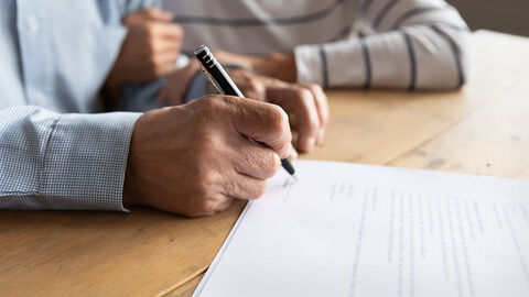 Person am Schreibtisch, die ein Dokument unterschreibt, dahinter eine weitere Person