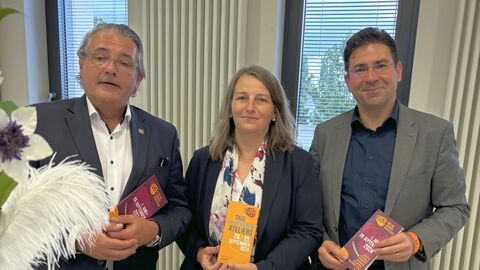 Zwei Männer und eine Frau an Tisch, mit Prospekt in der Hand