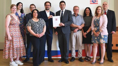 Fünf Frauen und fünf Männer stehen vor einer Leindwand für ein Gruppenfoto bereit. Zwei Männer in der Mitte halten gemeinsam einen Umschlag in den Händen.