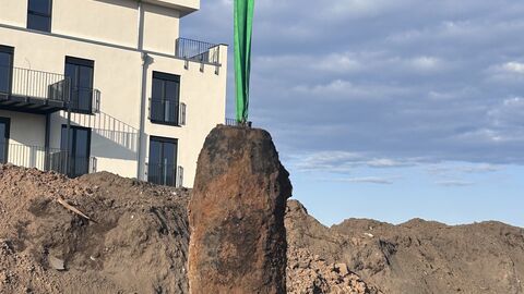 Bombe hängt an Baggerschaufel in der Höhe vor Gebäude