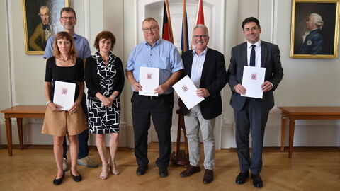 Zwei Frauen und vier Männer stehen vor einer Wand und blicken in Richtung der Kamera. Drei Männer rechts und eine Frau links halten Urkunden in ihren Händen. Im Hintergrund sind drei Fahnen und zwei Gemälde zu sehen.