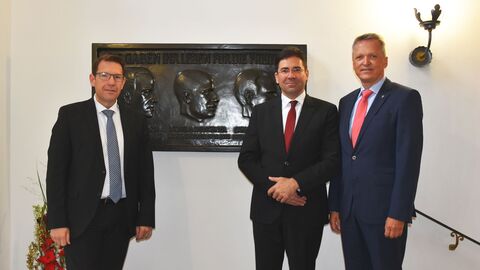Gedenkstunde Wilhelm Leuschner Drei Männer stehen in einem Treppenhaus vor einer Wandtafel und blicken in die Kamera. Die dunkel gestaltete Tafel zeigt drei Köpfe.