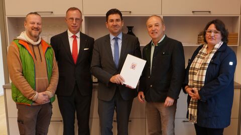 Fünf Personen, links vier Männer und rechts eine Frau, stehen nebeneinander und blicken in Richtung der Kamera. Der Mann in der Mitte hält eine Urkunde in den Händen.