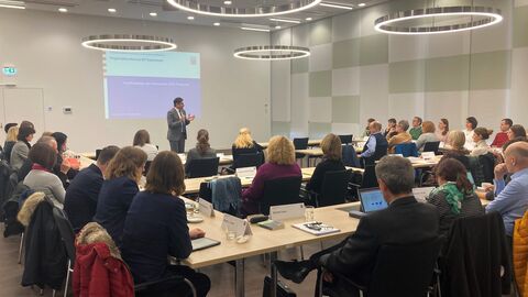 Zahlreiche Personen sitzen an einem großen viereckigen Tisch, vor Ihnen spricht jemand im Stehen