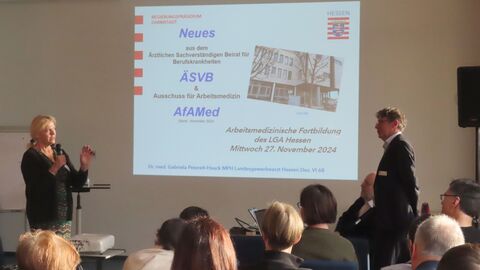 Fortbildung Landesgewerbearzt Eine Frau und ein Mann stehen sich gegenüber und schauen sich an. Im Hintergrund ist eine Präsentation auf eine Leinwand projiziert.