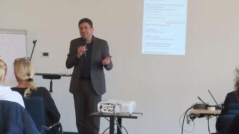 Fortbildung Landesgewerbearzt Ein Mann hält ein Mikrofon in seiner Hand. Er steht vor einem Publikum und spricht zu ihm.