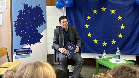 Ein Mann sitzt auf einem Stuhl und hält ein Buch in der Hand. Hinter ihm hängt eine Flagge der Europäischen Union. Vor ihm sitzen Kinder, deren Hinterköpfe zu sehen sind.