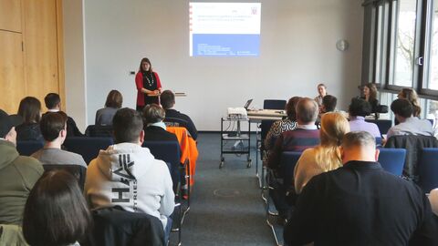 17 Menschen sitzen mit dem Rücken zur Kamera auf Stühlen. Sie blicken nach vorne. Dort hält eine Frau eine Präsentation.