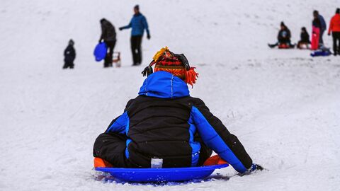 Ein Kind sitzt auf einem Schlitten und rutscht einen Hügel hinunter. Unten stehen weitere Menschen.