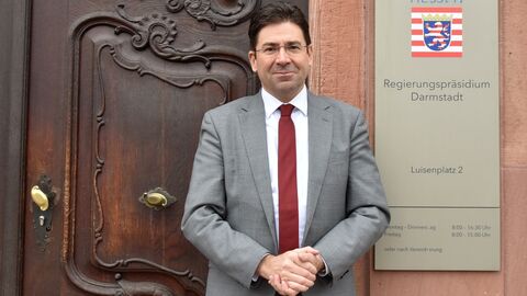 Ein Mann in einem Anzug steht vor einer großen Tür. Er blickt in die Kamera.