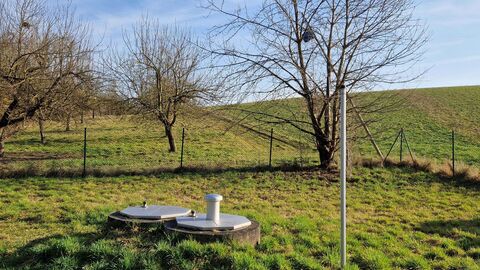 Brunnen Ober-Klingen mit eingezäunter Zone des Wasserschutzgebietes  Feldartige Wiese mit einigen wenigen Bäumen im Hintergrund und im Vordergrund zwei runde, silberne Deckel auf einem grünen Hügel