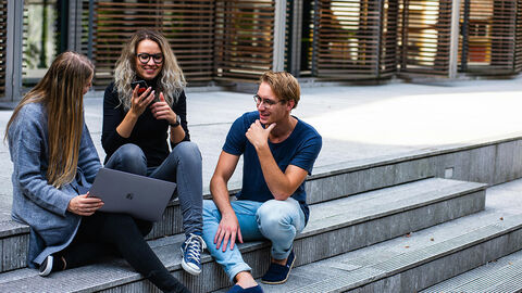 Drei junge Menschen sitzen mit Laptop und Handy auf Treppenstufen und unterhalten sich