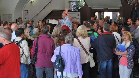 Eine große Besuchergruppe steht im Eingangsbereich eines historischen Gebäudes. In ihrer Mitte steht ein Mann, der ihnen vom Gebäude berichtet.