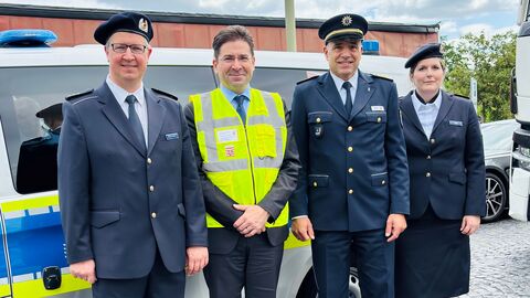 Drei Männer und eine Frau stehen vor einem Polizeiauto und schauen zur Kamera.