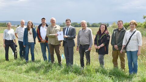 Zehn Menschen stehen nebeneinander auf einer Wiese und schauen zur Kamera. Die zwei Männer in der Mitte halten einen Zettel in den Händen. Im Hintergrund ist ein landwirtschaftliches Feld zu sehen.