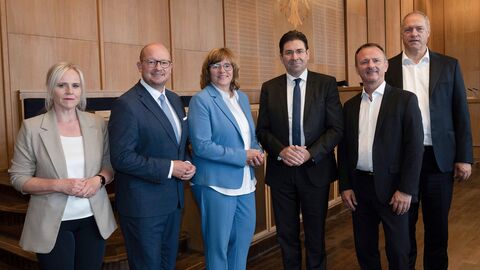 Zwei Frauen und vier Männer stehen nebeneinander in einer Reihe für ein Gruppenfoto bereits. Sie blicken alle in Richtung der Kamera.