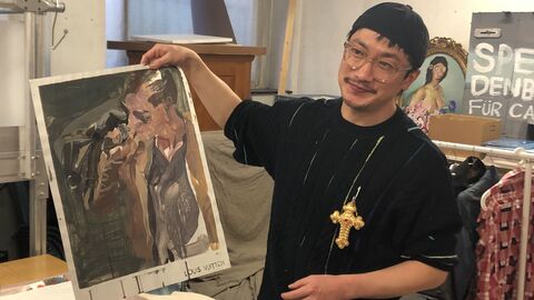 Künstler Liu Xue Ein Mann steht vor einem Tisch. In seiner rechten Hand hält er ein Bild hoch und präsentiert es.