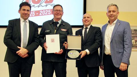 Vier Männer stehen für ein Gruppenfoto nebeneinander. Die zwei Männer in der Mitte halten eine Urkunde und eine Plakette vor sich.