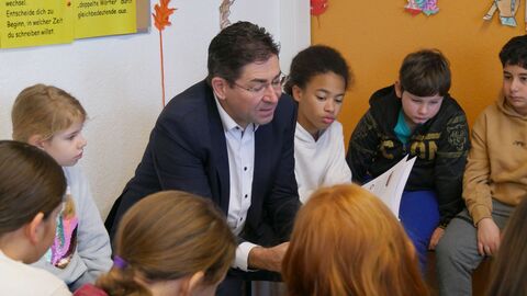 Ein Mann sitzt in einem Stuhlkreis mit Kindern und liest ihnen vor.