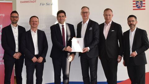 Sechs Männer stehen nebeneinander vor einer Foto-Leinwand für ein Gruppenbild in einer Reihe. In der Mitte halten zwei Männer gemeinsam eine Urkunde in ihren Händen.