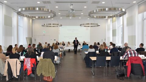 Personen sitzen in Saal, vorne spricht eine stehendende Person zu ihnen